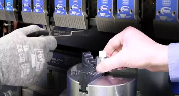 gloved hand holding bent metal in front of press brake and another hand inserting a piece into a grinder chuck