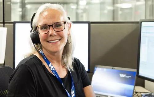 Mujer rubia con auriculares sentada frente a computadora sonriendo a la cámara